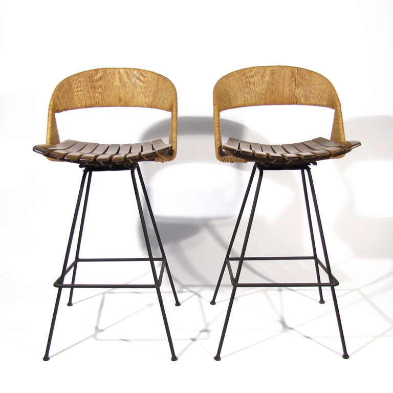 Handsome pair Arthur Umanoff swivel barstools. Very comfortable with swivel seats and curved raffia backs, bentwood slatted seats, both resting on satin black welded iron frames. Rare model with raffia extending down sides and tucking beneath seat.