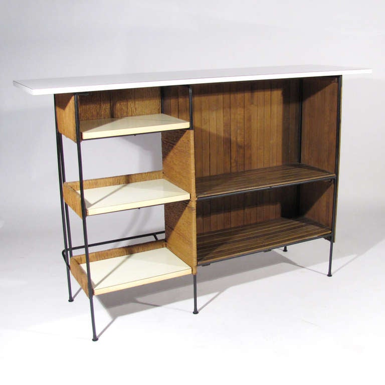 Arthur Umanoff for Raymor bar. Deep amber wood slats with white laminate top. Working side of bar has three white shelves with raffia fronts, and large shelved wood compartment. The consumption side (assuming the bartender is not nipping), comprises