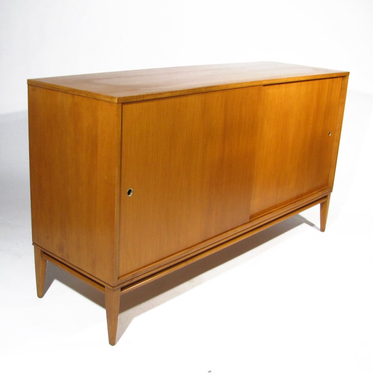 Paul McCobb Planner Group solid birch sideboard. Two doors slide to reveal one drawer with positionable shelf on the left, and three drawers above generous compartment on the right. Consistent deep honey color throughout entire piece. Foil label