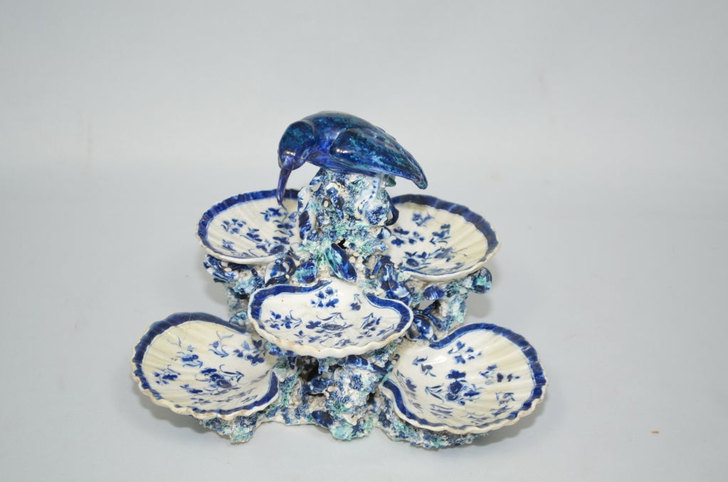Painted in blues and pale aqua with two tiers of scallop shell dishes that are decorated with a pattern of blue flowers. There are six shells in total, three on each level, which rest on a platform of coral surrounded by seaweed. The piece is topped