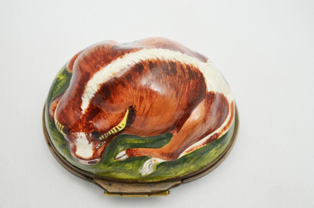 The top is shaped and decorated as a bull at rest on a small mound of grass. A pastoral scene featuring a tree, river, and country church is painted on the bottom of the snuff box. Hinge opens to reveal a white interior.