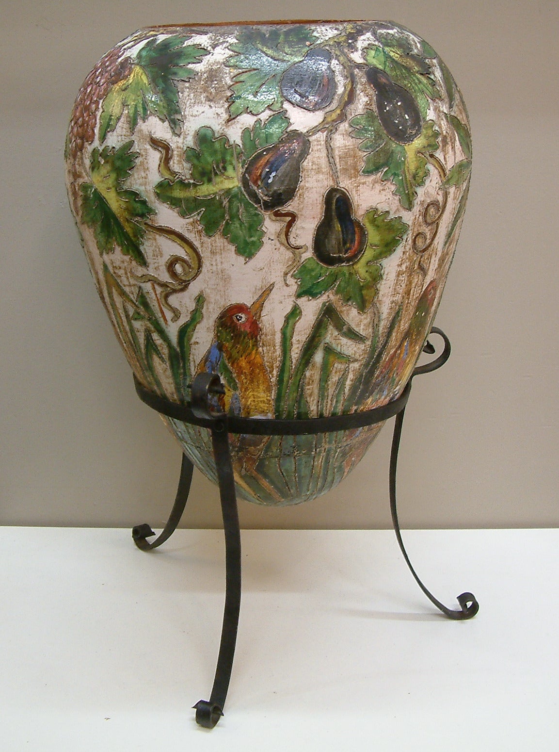 A very large and whimsical terracotta vase depicting birds, oranges, figs, pomegranates and grapes incised on a hi-glaze ground seated in a hand-forged and riveted iron frame.  Beautiful saturated color and detailing throughout.  The piece has an