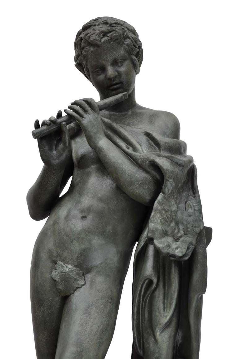 After the antique : a Ducel Foundry cast iron figure the Faun with Pipes, French, circa 1870, standing naked except for a fig leaf and playing a flute, next to a pedestal draped with a lion skin, on rectangular base (recent bronze patina). The Faun
