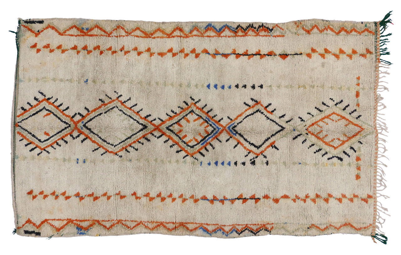 This eclectic vintage Moroccan Azilal rug features a stacked diamond protection design along the center with colors of orange, black, blue and a muted mint green on a creamy beige field. Azilal rugs with their plush pile and sturdy weave make them