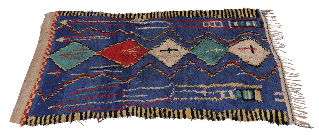 This eclectic Moroccan rug features contemporary abstract geometrical designs with a colorful palette of ivory, white, red and turquoise hand-knotted in a modest tribal pattern on a blue field. It is enclosed in a beautifully contrasting yellow and