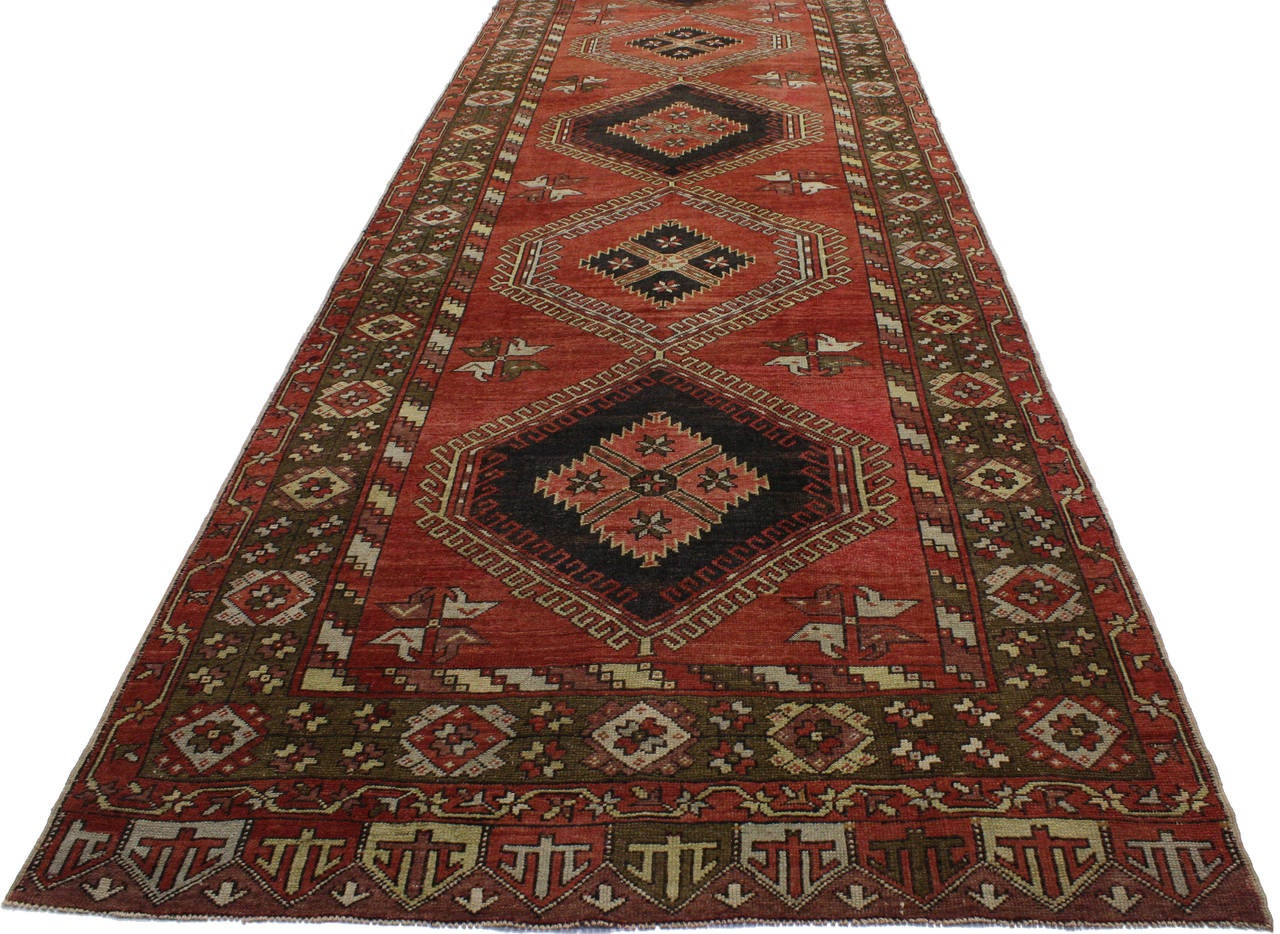 Full of character, stately presence and Mid-Century Modern style, this vintage Turkish Oushak carpet runner showcases an extravagant geometric design and grand scale motifs rendered in a vibrant color combination of red, olive green, brown with