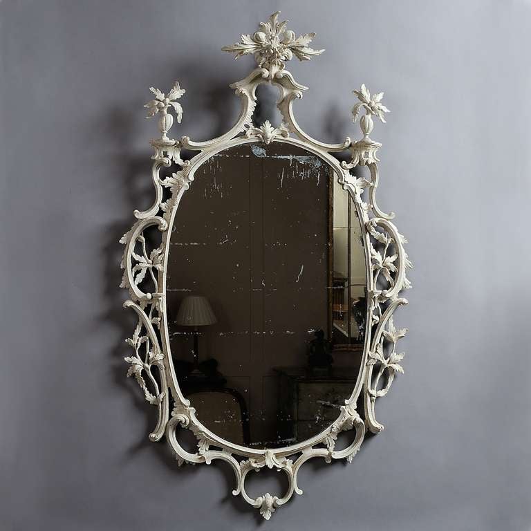 A FINE GEORGE III ROCOCO WHITE-PAINTED OVAL MIRROR, CIRCA 1760.
The delicate frame carved in deep relief with c-scrolls and foliage.  The pediment cresting surmounted by a spray of fruit and foliage and flanked by asymmetrical urns on rockwork