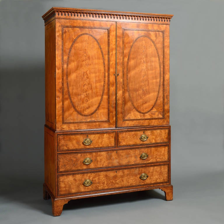 A fine George III satinwood clothes press, circa 1790.
Banded throughout in Kingwood and inlaid with ebony and boxwood strings, the cornice with gothic arcading and pendant acorn finials, the oval panelled doors enclosing six mahogany-lined slides.