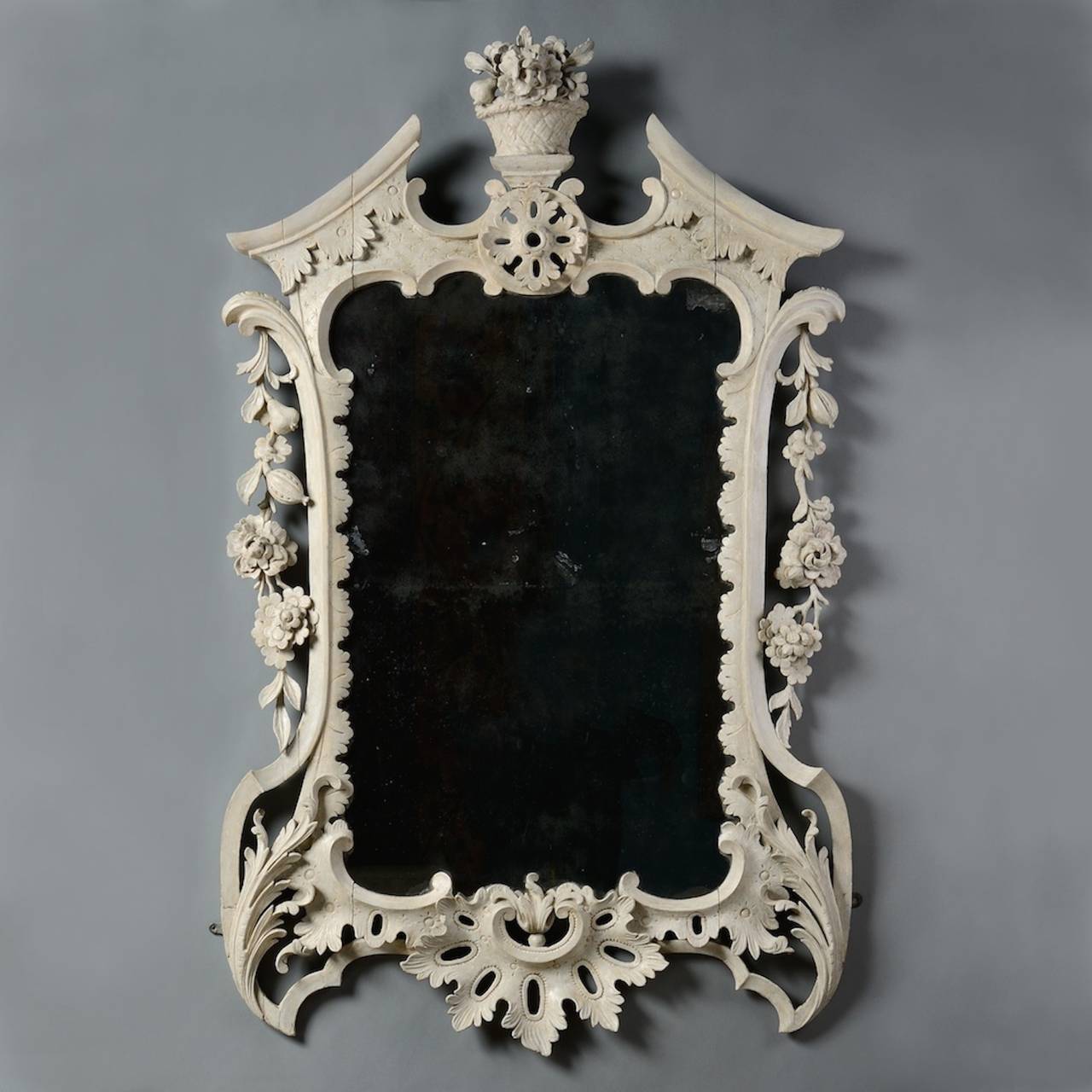 An unusual George II white painted mirror, original plate and decoration, circa 1750.
Carved with scrolls and foliage, the pediment cresting centered by a basket of flowers, the sides hung with festoons of fruit and flowers.