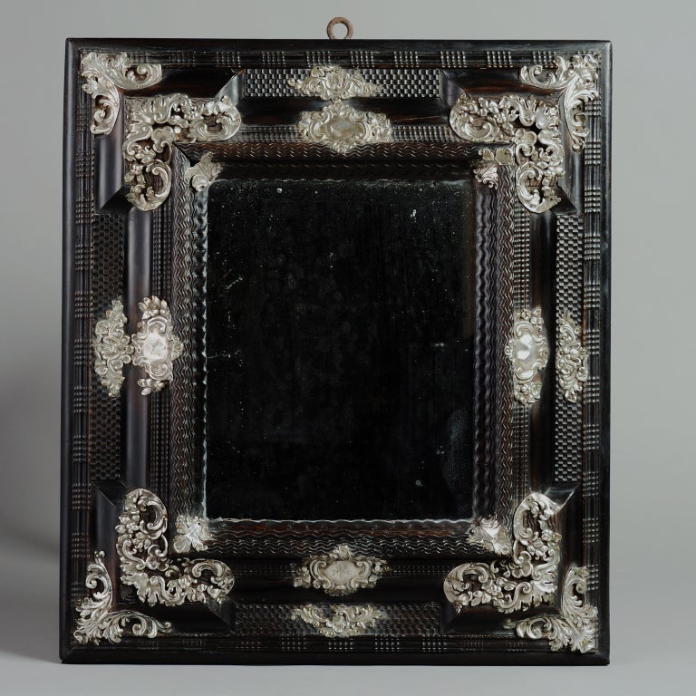 A RARE FLEMISH BAROQUE SILVER-MOUNTED EBONISED MIRROR, ANTWERP, CIRCA 1680.
The engine-turned ripple moulded eared frame applied with repousse silver foliate scrolls and cartouche panels, one engraved with the initials S G and a coat of arms, some