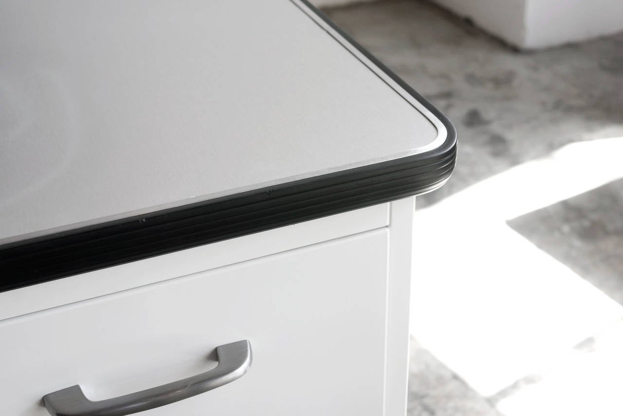 This unique, sized-down tanker desk features a single pedestal with locking file drawers. Bumper top with brushed steel Formica surface. Newly refinished in a white powder coat. The look of a mid century tanker with a minimal, console-style profile.