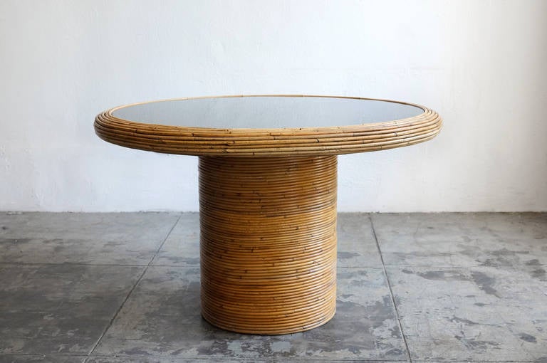 A wonderfully unique rattan dining table by Henry Olko for Willow and Reed, signed and dated 1978. Black glass sits atop a thick cylindrical base.The rattan is in very good condition. The glass table top is intact with no chips; light scuffing.  
A