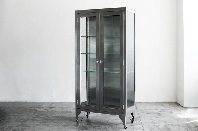 Stunning 1960s medical instrument and supply cabinet. Composed of stainless steel with glass on three sides. Features three wired glass shelves with locking glass double doors. Stands off the ground on tapered leg and rolls on casters. 

In the