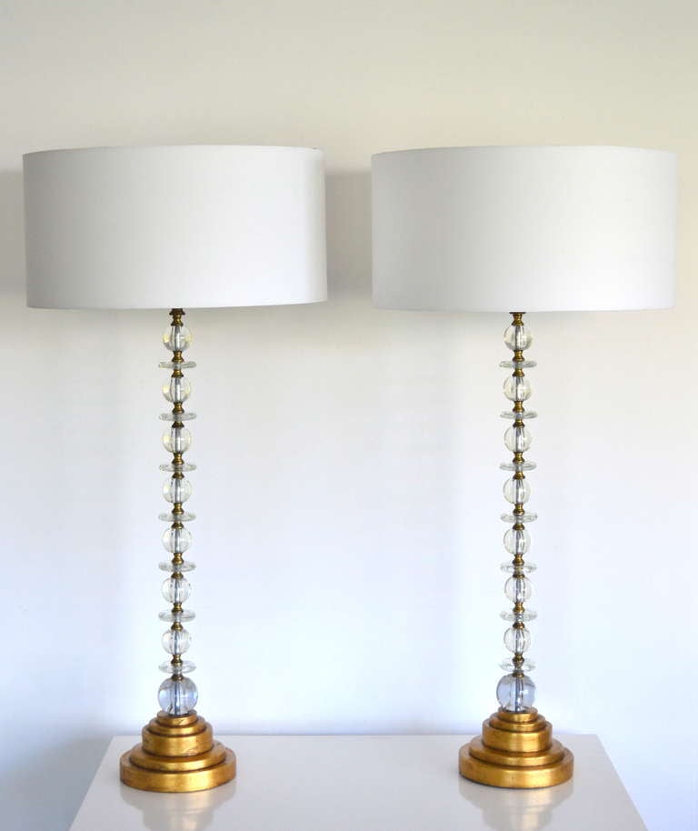 Glamorous pair of tall Mid-Century Modern stacked glass table lamps, c. 1940s - 1950s. These deco-inspired clear glass lamps in the manner of James Mont are composed of alternating blown glass and brass elements and are mounted on turned gilt wood