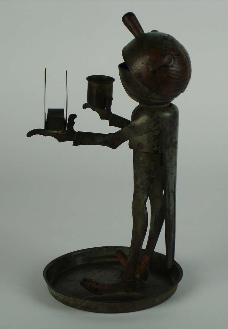 A hammered wrought iron,pressed steel & brass tobacco necessary representing a servant.The head being an ashtray,one hand holding a candlestick,the other holding a matchbox support.This item was originally painted in red & blue.
