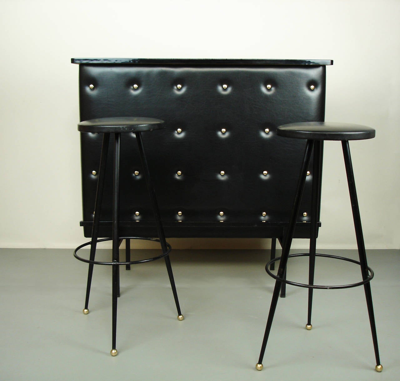 A bar with a in black laminated top and an enamelled metal support, upholstered with tufted vinyl and bronze buttons.
Two stools with black enamelled metal feet and vinyl seats. Height: 30.25 in.