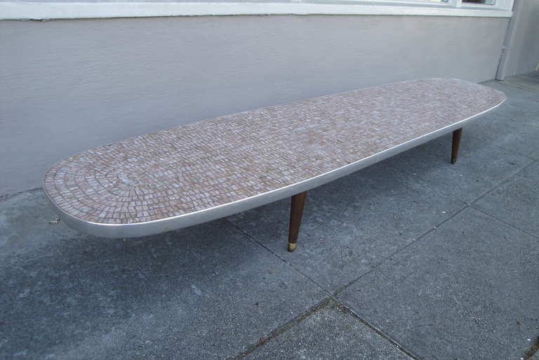 Long and low, mosaic tile surfboard coffee table with peg legs. Tiles have an unusual mica or copper content that is evident upon close inspection. Peg legs attach with threaded hardware and can easily be replaced with replacement legs to change