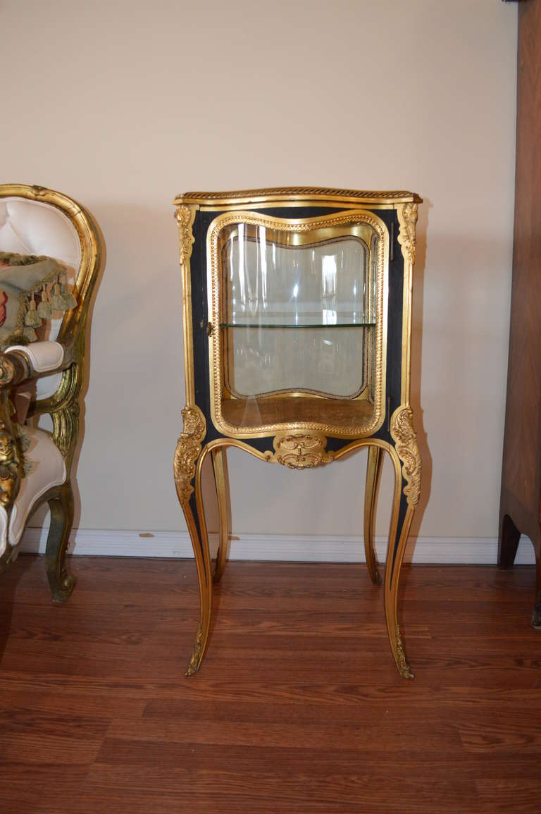 A jewel of a small Louis XV style vitrine with extensive carved bronze details.
The top has a beveled glass, the side glasses are curved and there is one glass shelf. The bottom shelf is cover in a velvet fabric and there is a silk cord all around