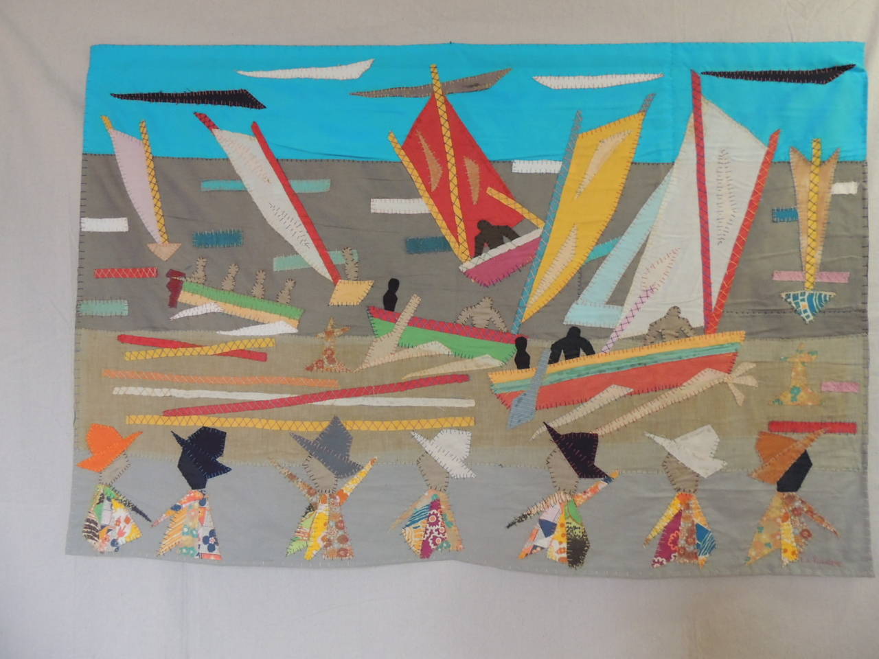 Applique patchwork quilt wall hanging art by a Bahamian artist. Applique figures of fisher men arriving at fishing port greeted by villagers. The bright colors of the boats and sails. In shades of red, blues, yellow, orange. The villages clothing is