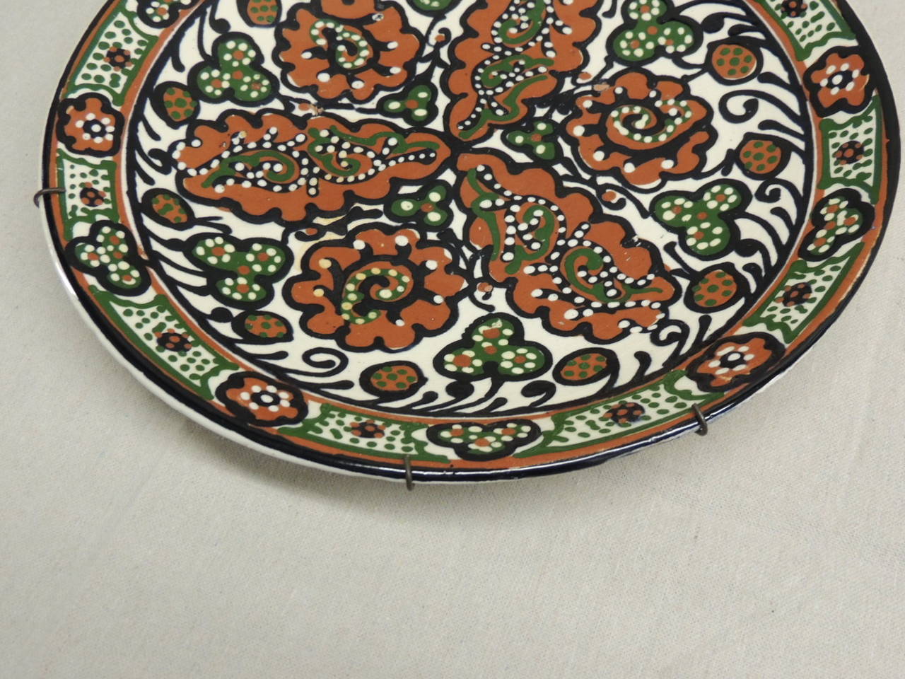 Large glazed ceramic painted plate/charger. Depicting flowers and leaves in shades of orange, green and black. Hanging wires on back.