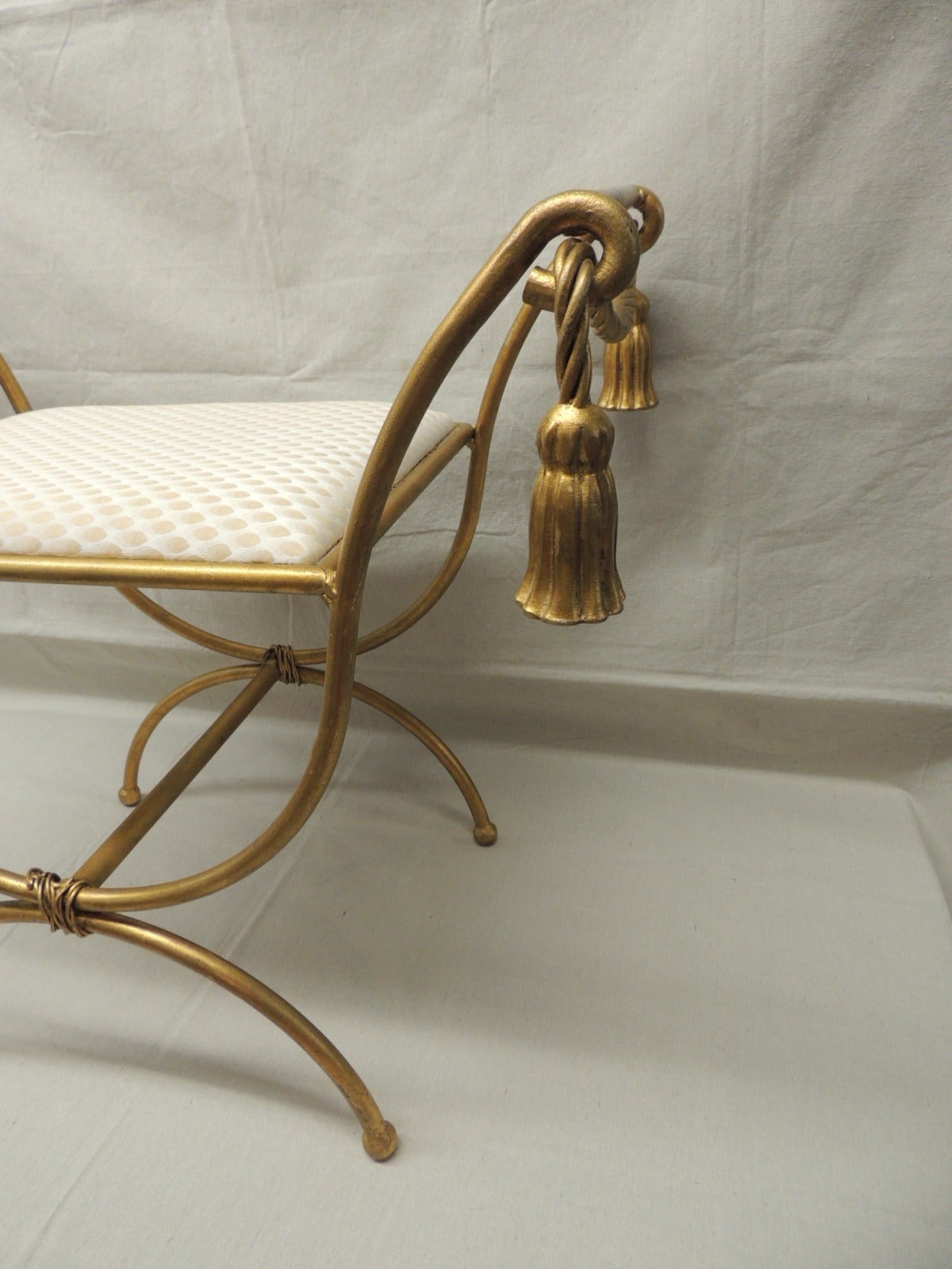 Gilded rope and tassel vanity stool or bench. X-base and upholstered in a silk fabric. Fabric has a small run on corner, but seat unscrew for easy reupholstering.