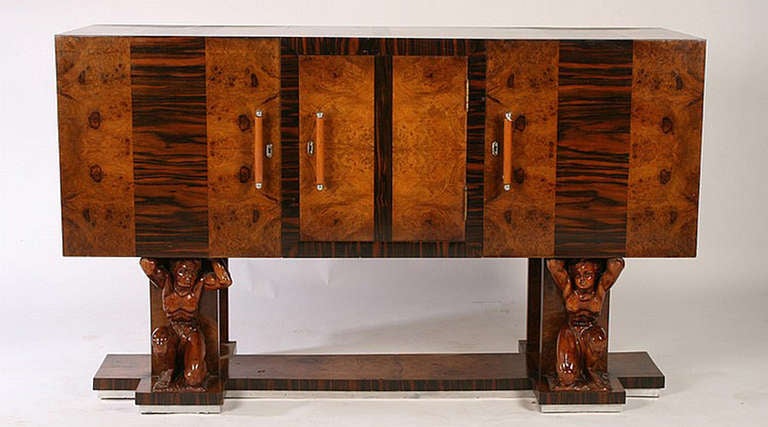 This double pedistal Deco piece is in great condition. Burled walnut and rosewood veneer with metal trim. Columns are being held up by a carved man, perhaps Atlas. Rosewood and metal trim around the very base. Three doors with bakelite and chromed
