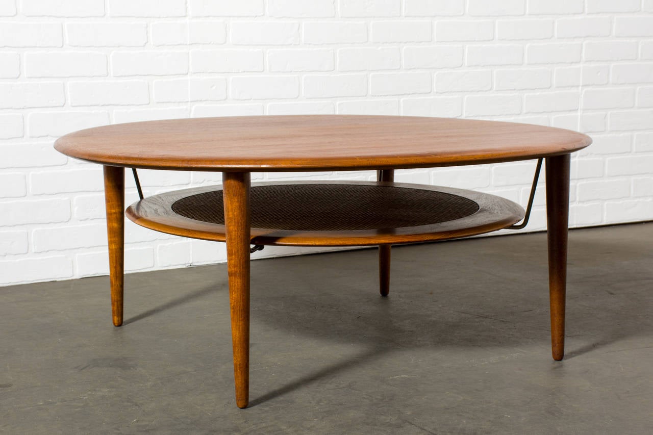This is a vintage Mid-Century round solid teak coffee table designed by Peter Hvidt & Orla Molgaard Nielsen for France & Sons in the 1950s, Denmark (model FD 515). It has a cane shelf below that is suspended by brass brackets.