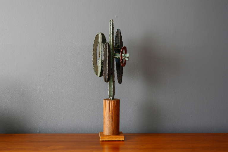 This is a vintage Mid-Century brutalist cactus sculpture.  The metal cactus has a green tone with red and blue details. It rests on a heavy wood base.