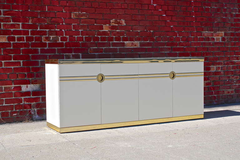This vintage sideboard was designed by Pierre Cardin for Dillingham in 1978. It is high gloss ivory laminate with polished brass trim and has four doors that conceal two adjustable shelves and two drawers.