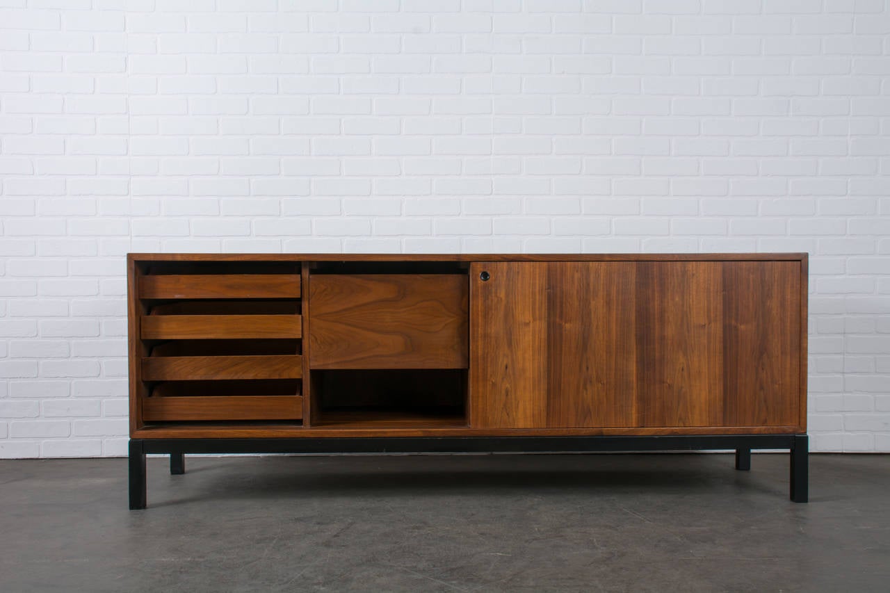 This is a Mid-Century Modern walnut cabinet that was designed by Jacques S. Guillon. It has two sliding doors, the right side conceals an open storage space and the left side conceals four medium sized drawers and one large drawer. This storage