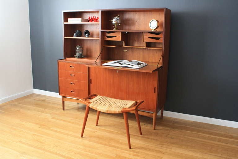 This is a Mid-Century Modern teak storage piece with sliding doors on the bottom right that conceal one adjustable shelf, four drawers on the bottom left, a drop leaf desk on the upper right, and two adjustable shelves on the top left.