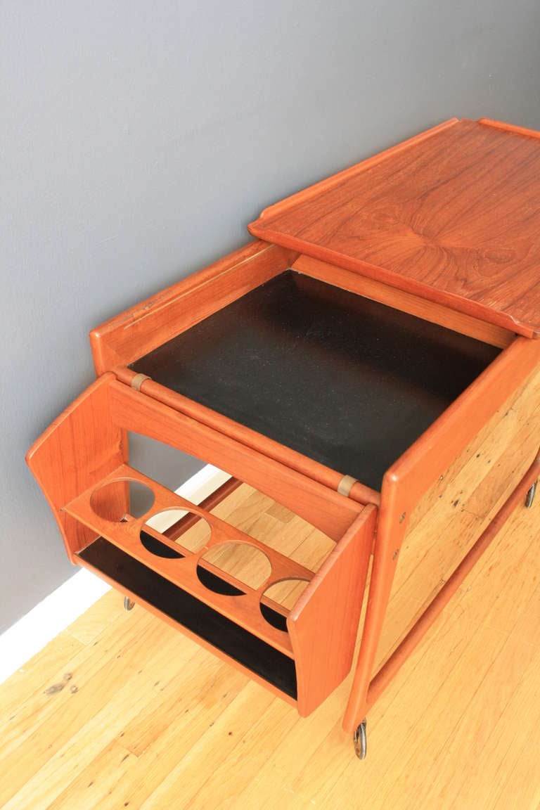 This is a vintage Mid-Century teak bar/cocktail cart by J. Ingvard Jensen. It has a hanging bottle holder that is removable and is stamped DESIGN J. INGVARD JENSEN, ODENSE, MADE IN DENMARK. A sliding top reveals an interior compartment.