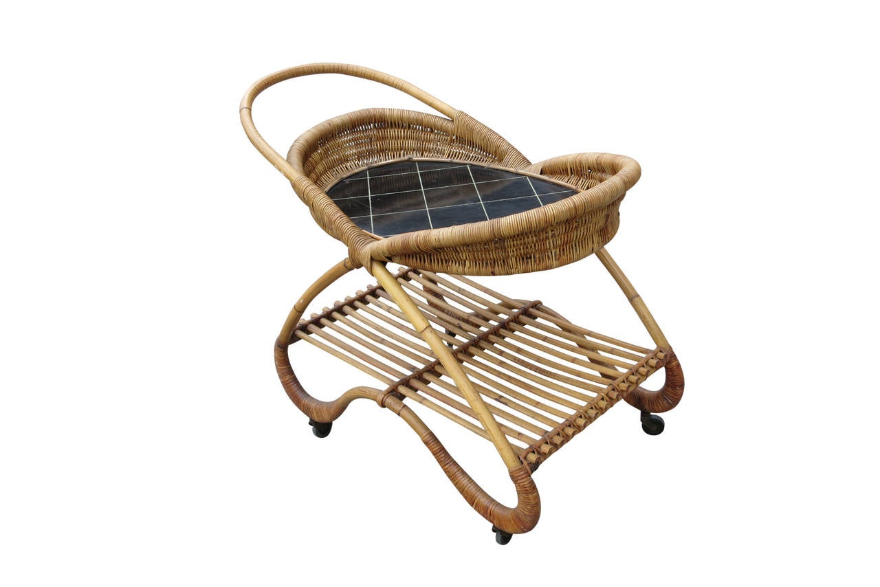 This unique 1950s bar cart features a free-form designed frame with two shelves. The lower shelf is made of rattan slats and the top shelf made with a black tile top sitting in a woven wicker basket.

The cart easily rolls on four