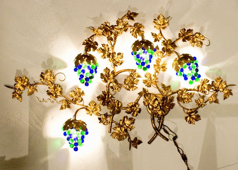 This wall sconce, in the organic shape of a sprawling grapevine, was made in Italy and features four light-up Murano glass grape clusters mounted in a large gilt metal frame. The cord is intertwined with metal chain and adorned with golden leaves.