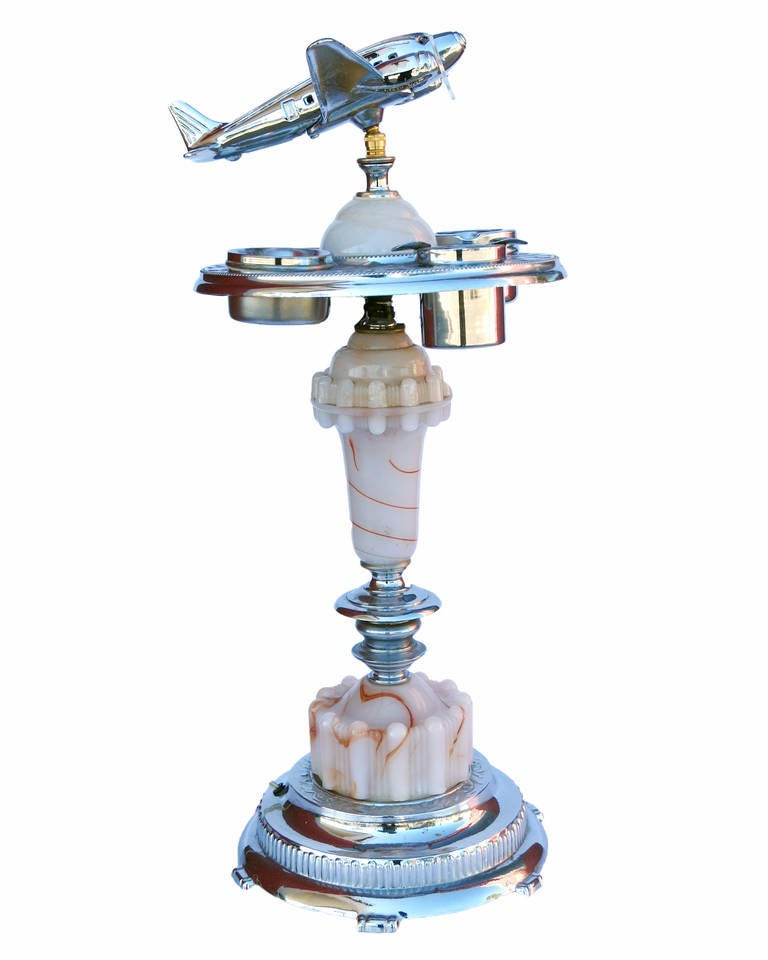 Chrome Art Deco ashtray stand featuring a decorative light up airplane on top with a glass light up bass on the bottom.

The stand comes with a built in light with 3 ashtrays and 1 tobacco holder.
This was used in the day as a conversation for