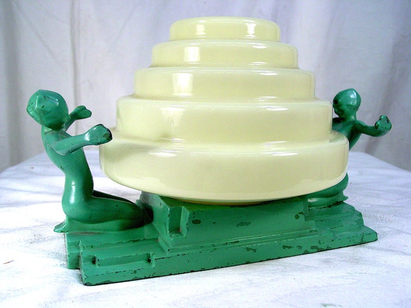 This evocative 1920s Frankart table lamp depicts a Classic Art Deco motif of two nude figures embracing a central tiered shade in cream toned glass. The base and green painted finish are original to the piece. 
The base is stamped “Frankart Inc.,”