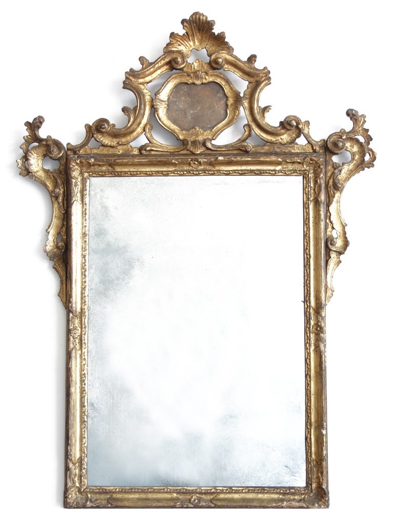 The gilt carving rises up to a rocaille and foliate crest surrounding a small glass plate with the faintest memory of its silver. The larger silvered glass has a wonderfully atmospheric cloudiness, and is surrounded by scrolled pendants along the