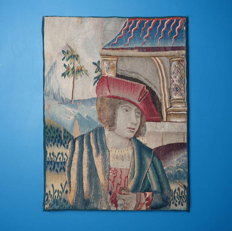 This charming tapestry of a young man standing before a building with a hilly landscape in the distance is undoubtedly a fragment of a much larger work. His richly decorated clothing, with light playing off the luxurious surfaces and deep folds, is