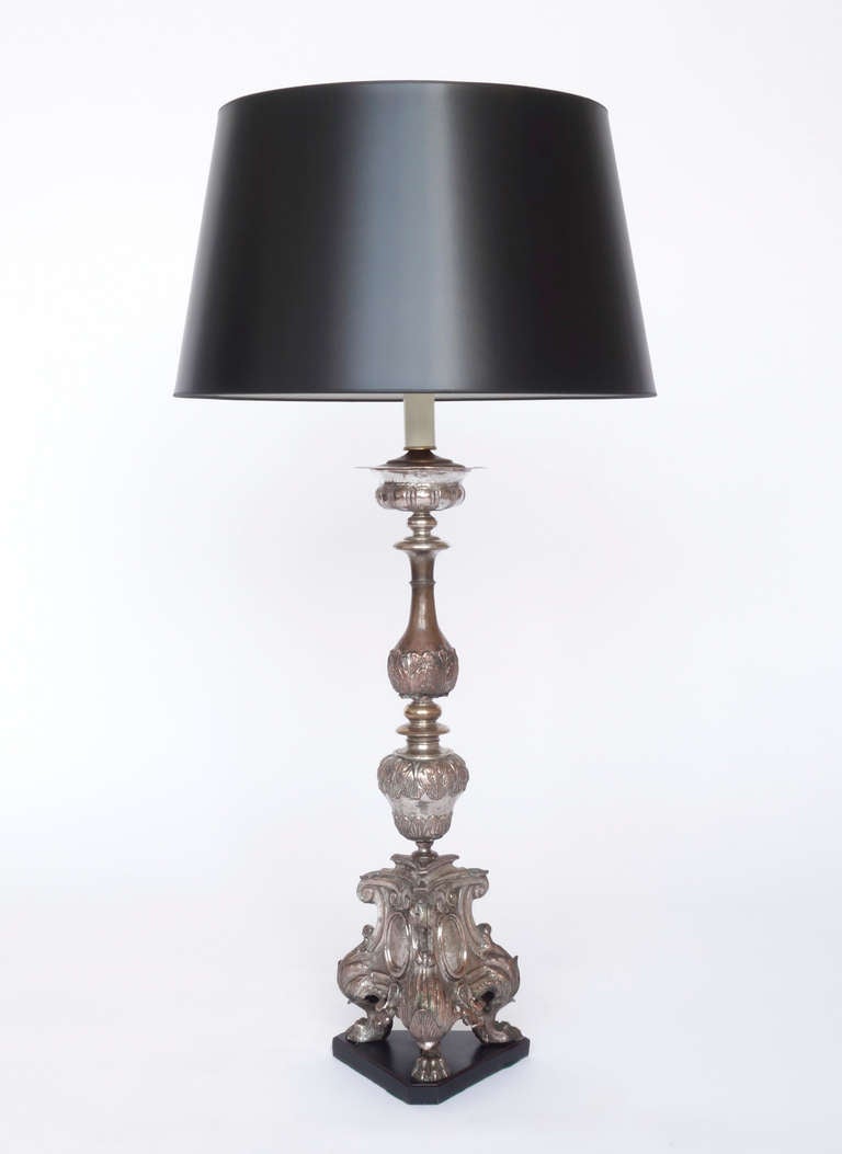 A pair of Italian silvered copper repoussé altar prickets fitted as table lamps. Baluster stem with paw feet and rocaille decoration surrounding an engraved cartouche. The base is three-sided. Two of the cartouches bear inscriptions: 