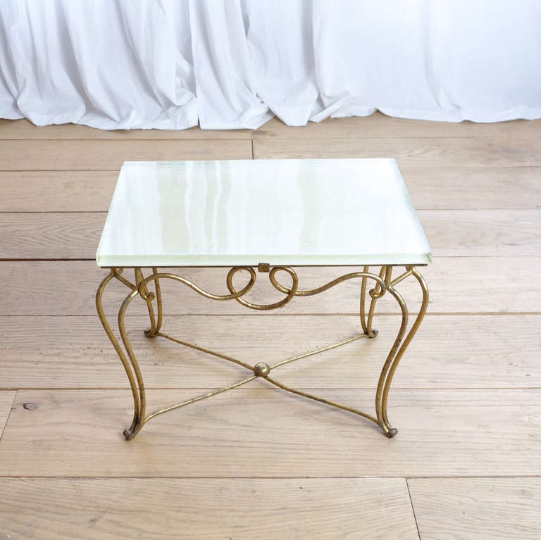 The loops and curls of this gilt iron table base are typical of French Art Moderne designer René Prou and his contemporaries. We enjoy the contrast between the gilt arabesques and the icy rectangle of the thick glass top.