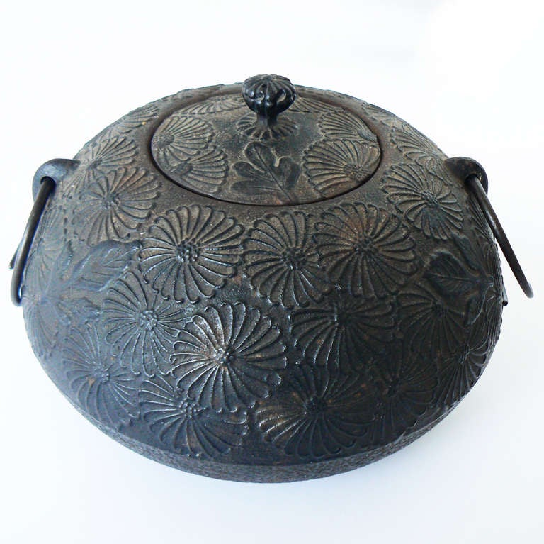 This Meiji-period iron chagama has a wonderful gravity. It is decorated with stylized chrysanthemums. The loop handles are typical for this type of pot, which is used to heat the water for tea ceremonies. Seen in situ resting atop a Jean Pascaud