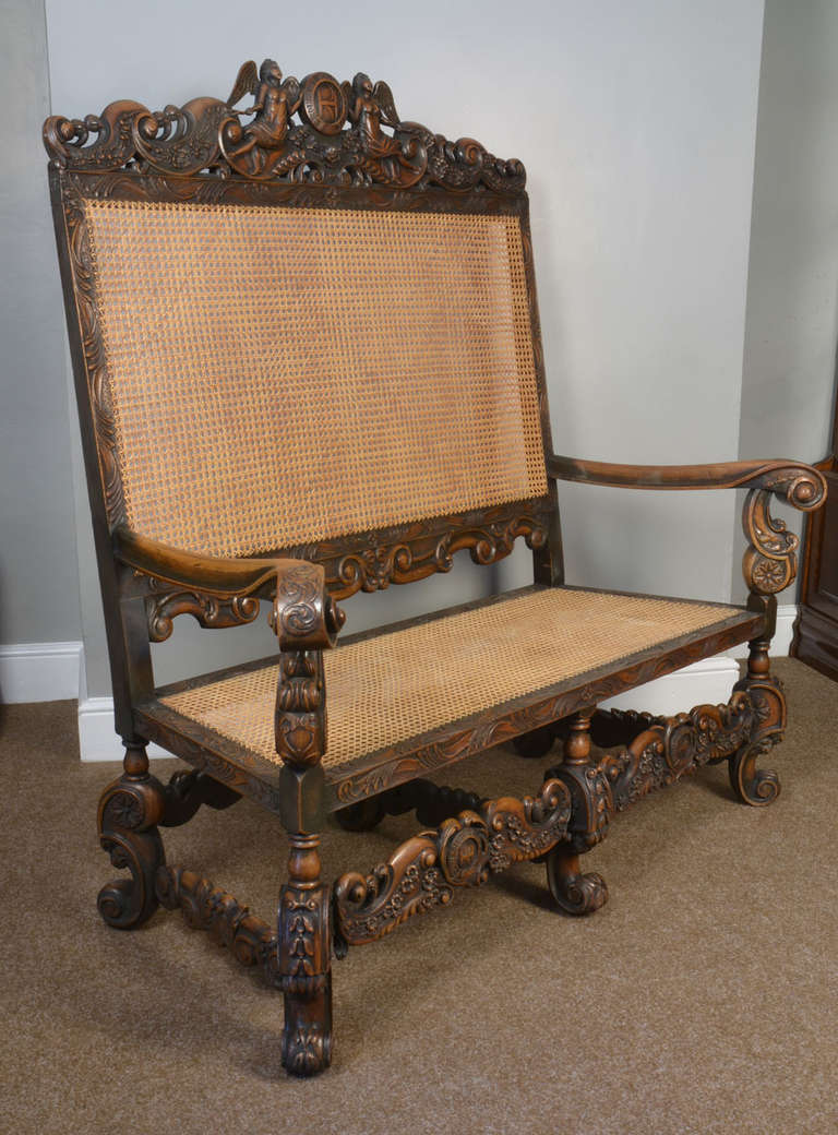 Walnut high back settee in the late 17th century style. The rectangular cane work back beneath an elaborately carved cresting centered by a cartouche bearing the initials 'RH' within the garter, flanked by two winged figures and floral-carved and