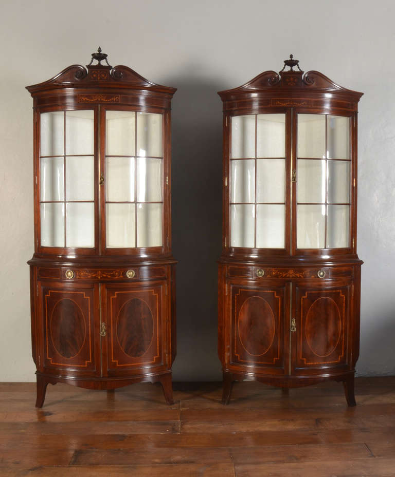 pair of mahogany bow fronted corner display cabinets by Shapland and Petter, inlaid with boxwood stringing, the upper part with carved urn finial and moulded cornice above inlaid panel of floral swags. The glazed bow fronted top section fitted with