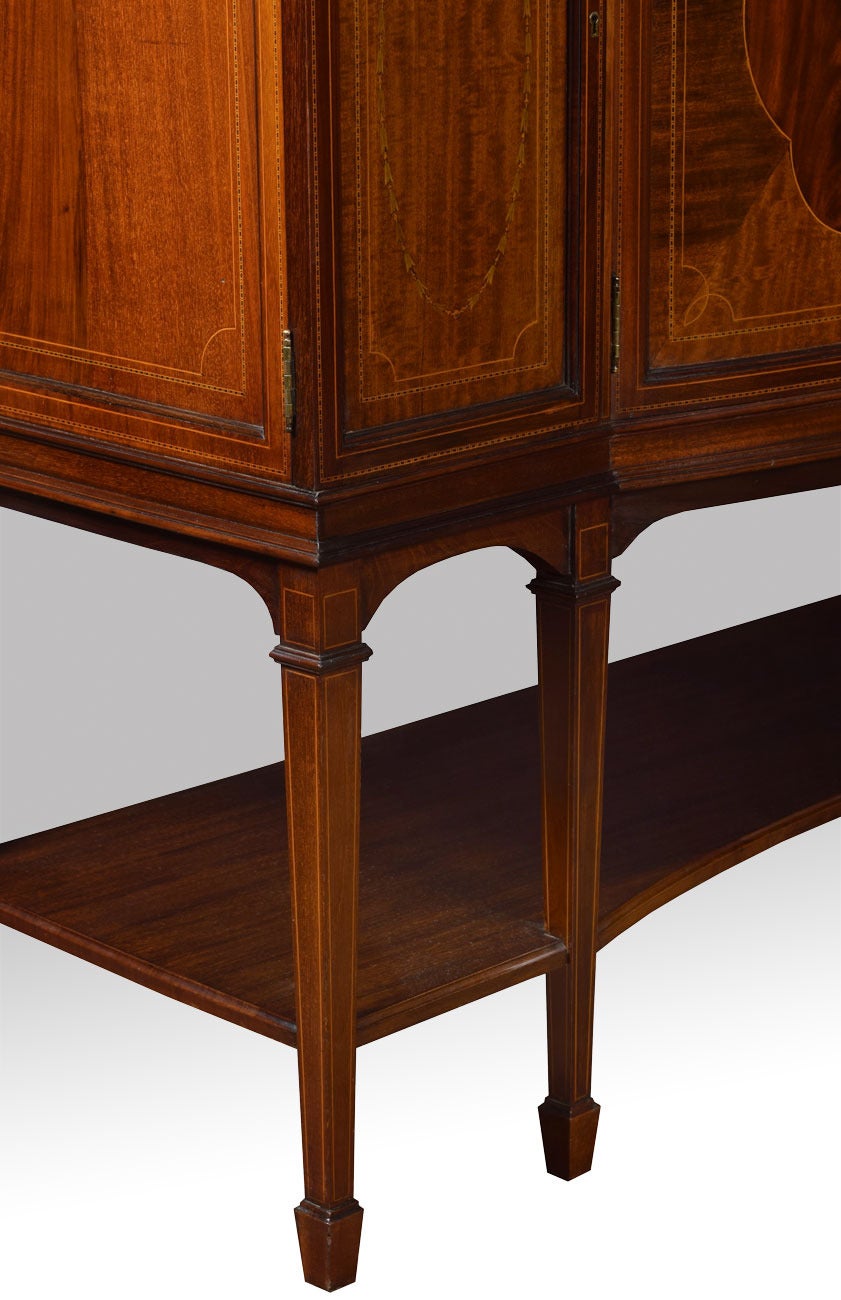 Edwardian Mahogany inlaid display cabinet of bow breakfront form, the moulded cornice and checker strung freeze, over a pair of conforming curved glazed doors enclosing two glazed shelves, the base section having curved cupboards enclosing shelved
