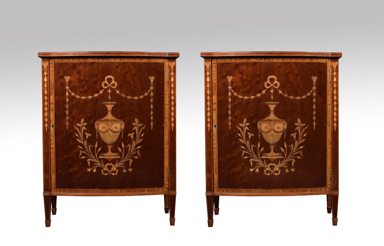 Pair of mahogany and inlaid cabinets, the bow fronted inlaid tops above long single doors inlaid with large central urns, bell flowers and ram's heads, having satinwood crossbanding throughout, the door opening to reveal shelved interior all raised