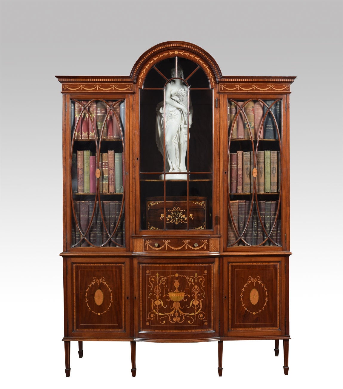 Late 19th century Sheraton Revival mahogany inlaid display cabinet bookcase, the dome top with dentil-inlaid cornice with bell-flower, above three astragal-glazed doors enclosing wooden shelves, the base section fitted with central slender drawer