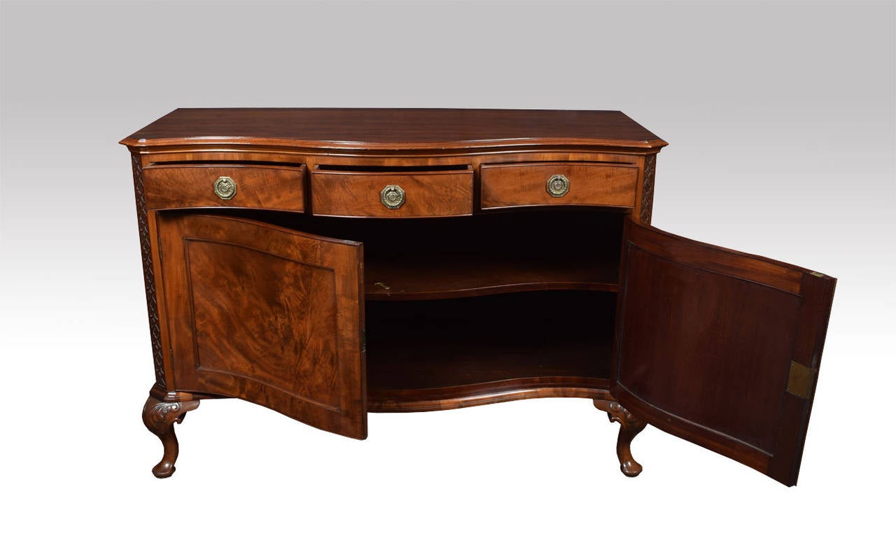 Mahogany serpentine fronted sideboard the shaped top above three freeze draws with brass tooled handles to the base section fitted with two mahogany panelled doors opening to reveal large storage area with single shelf, all raised up on leaf capped