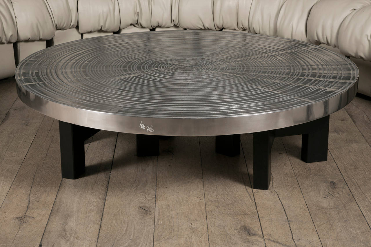 Round coffee table in cast aluminium supported by three black steel tripod legs. 

The textured top surface with circular graduating rings was inspired to the artist by drop of water.

Signature engraved by the hand of the Belgian artist on the