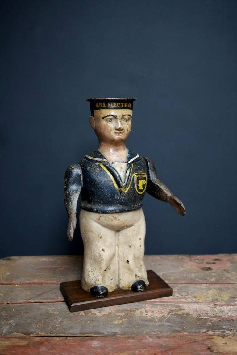 A cast iron figure of a British Navy Seaman. 
Cap marked HMS Electra. 
We think this was part of a donation box for the war effort. 
His lapel pocket has a slot for the donation and then we assume you pushed his right arm that is spring loaded,