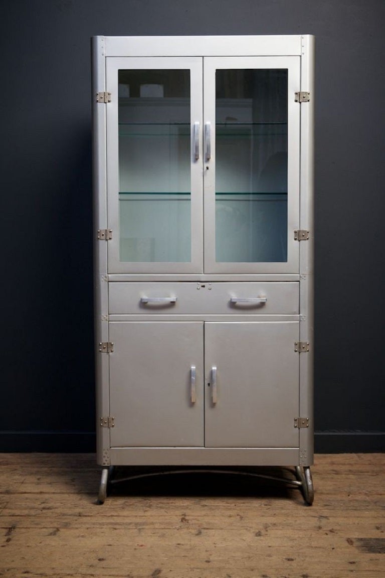 Grey enamel glazed medical cabinet. 

Glazed double doors with original lock above a single drawer and a pair of lockable solid doors. 

Tubular legs, alloy handles, glass shelves, bronze locks. 

English 1960s. 

H:176 W: 82 D: 37 CM